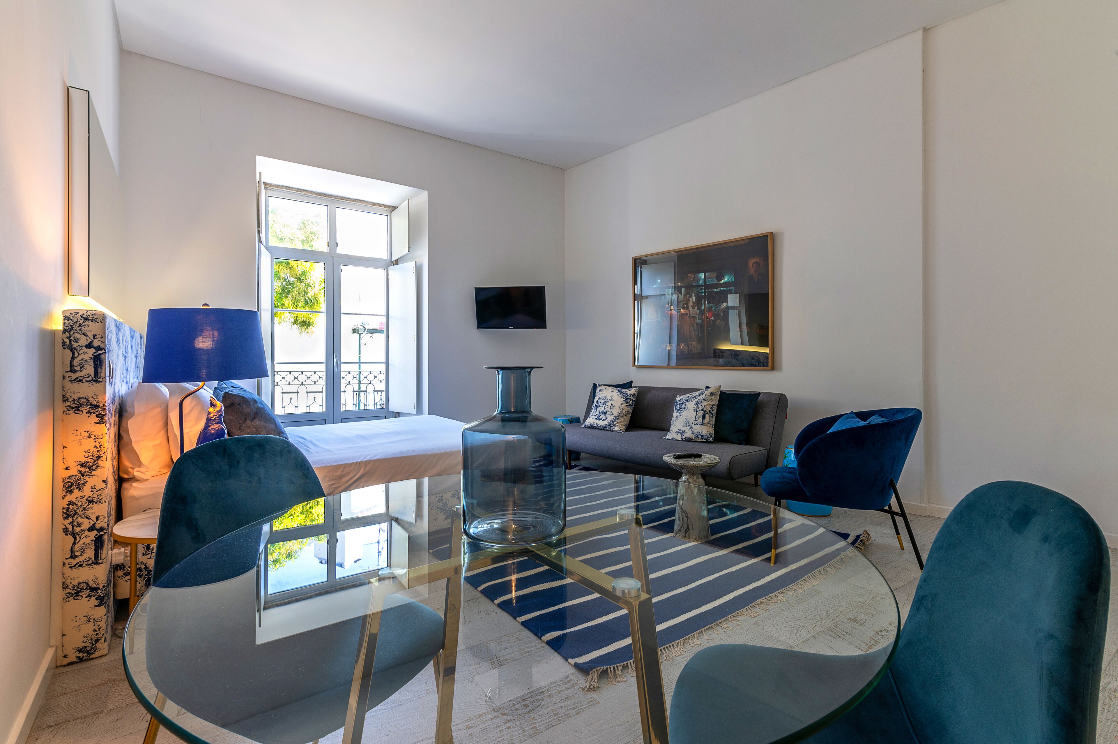 A modern living room with a glass coffee table, blue chairs, a sofa, and a TV mounted on the wall.