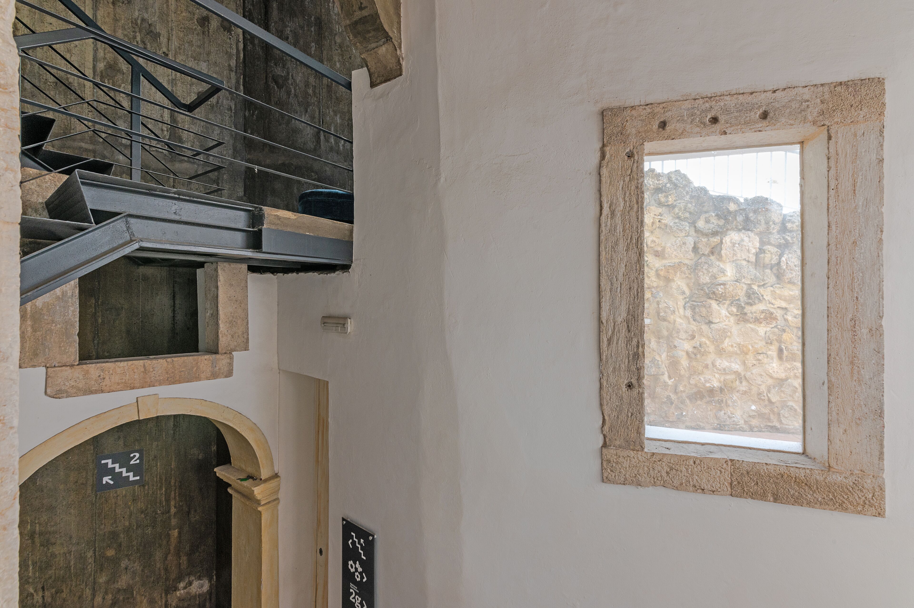 Un escalier avec une rampe en métal, une fenêtre avec un cadre en pierre et une porte sur laquelle est apposée une indication signalant la direction du deuxième étage.
