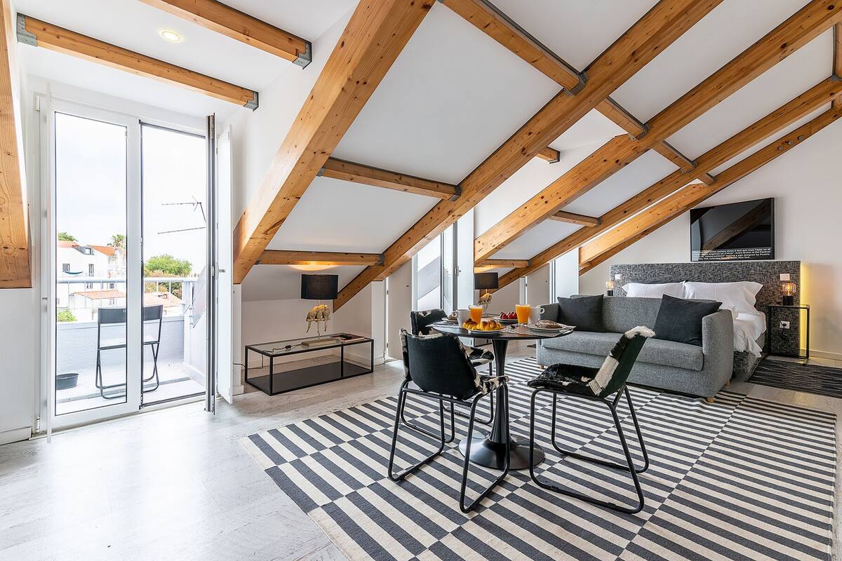 A modern loft bedroom with exposed wooden beams, a large window, a cozy seating area with a sofa and chairs, and a dining table with breakfast items.
