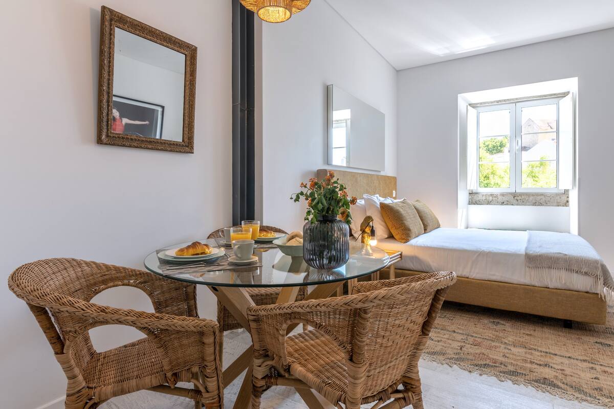 A room with a glass dining table, wicker chairs, a bed with a white headboard, a large mirror, and a window with a view of greenery.
