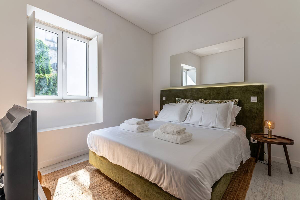 A bedroom with a bed, a window, a mirror, and a bedside table with a lamp.