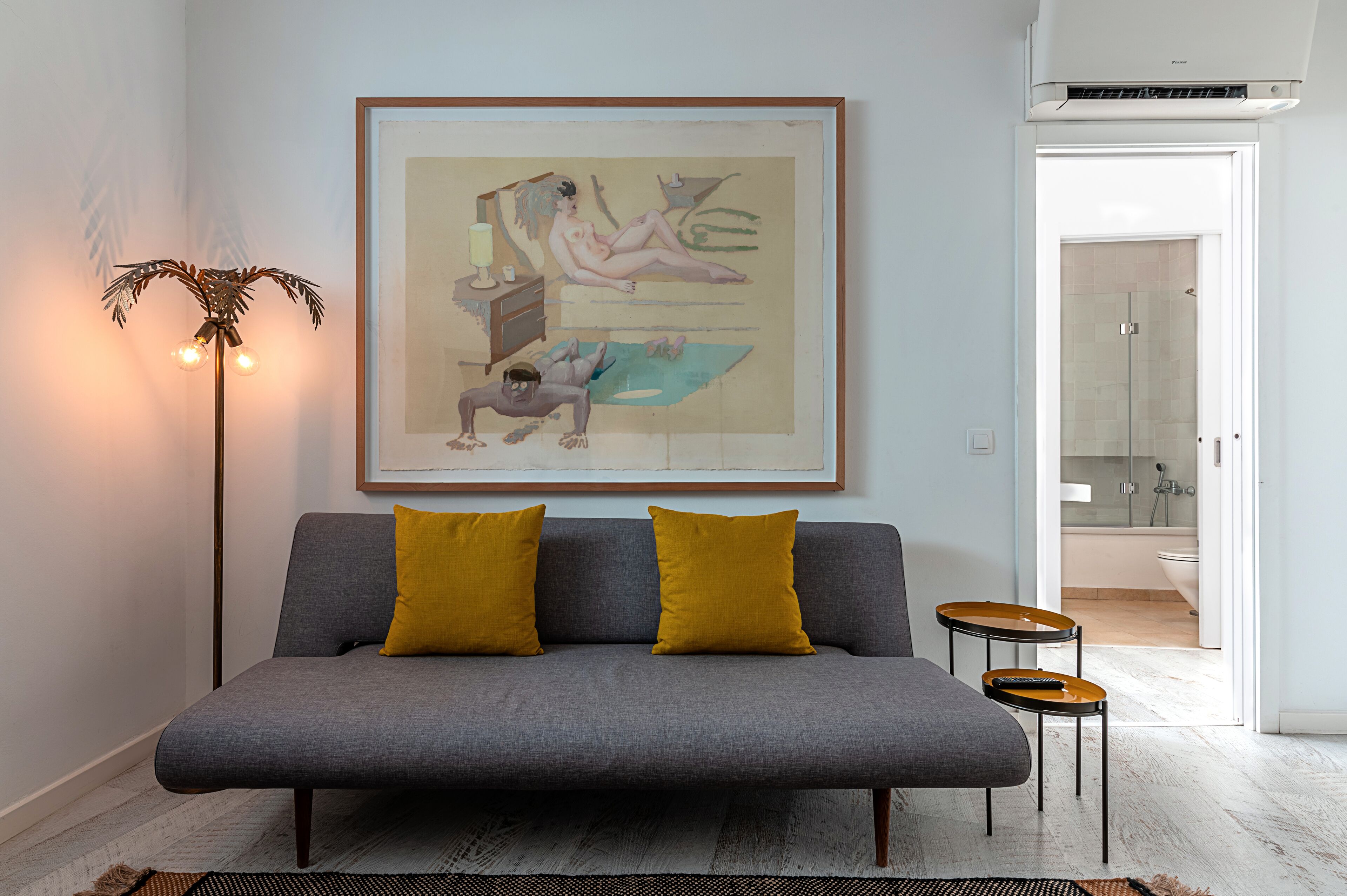 Een moderne woonkamer met een grijze bank, twee mosterdgele kussens, een lijst met abstract schilderij, een vloerlamp en een klein tafeltje met een afstandsbediening.