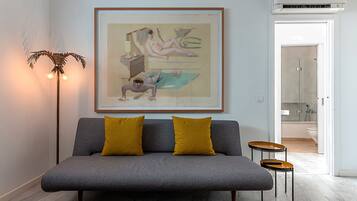 Een moderne woonkamer met een grijze bank, twee mosterdgele kussens, een lijst met abstract schilderij, een vloerlamp en een klein tafeltje met een afstandsbediening.