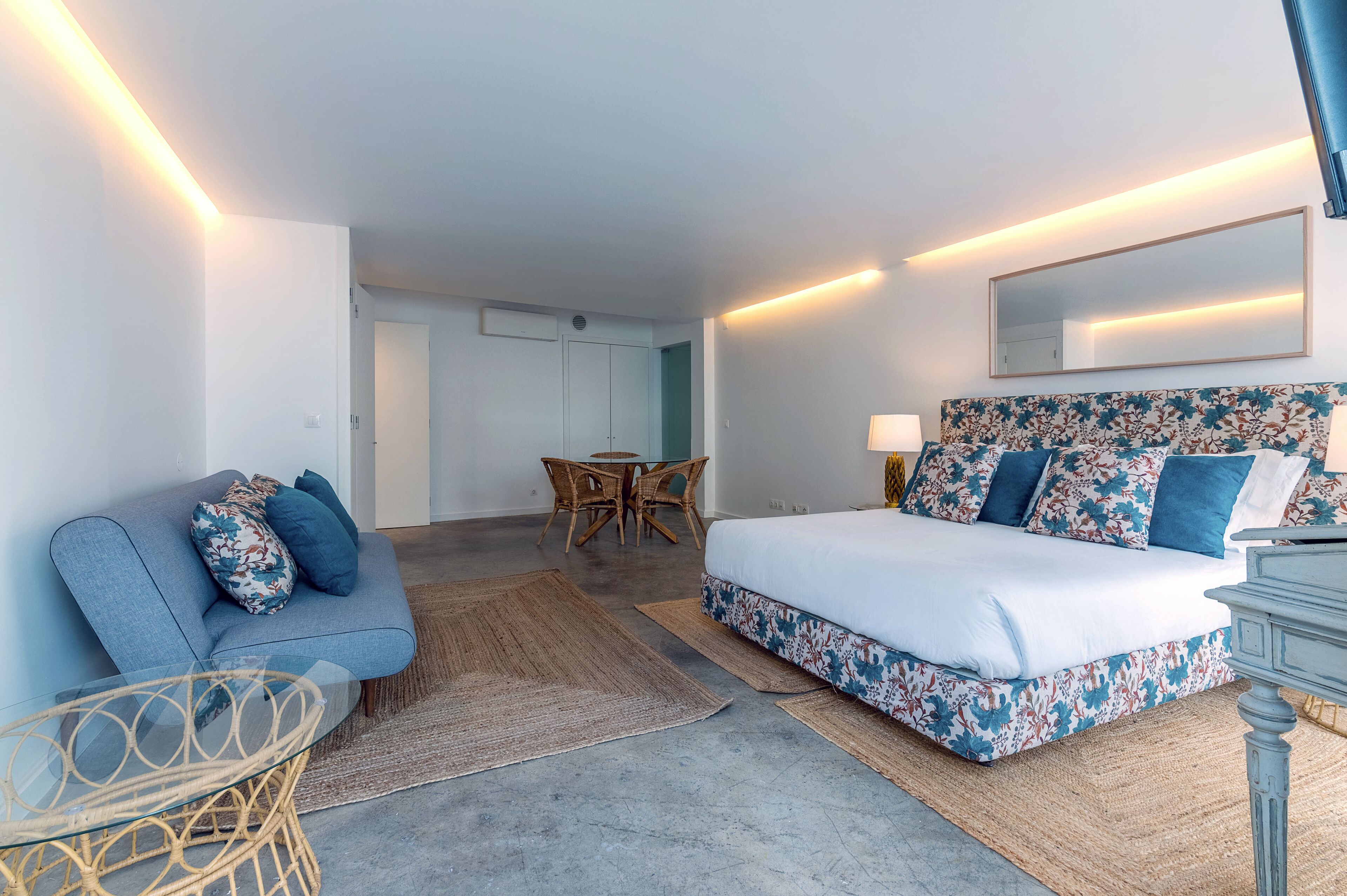 A modern hotel room with a large bed, a sofa, a dining table with chairs, and a small table with a lamp.