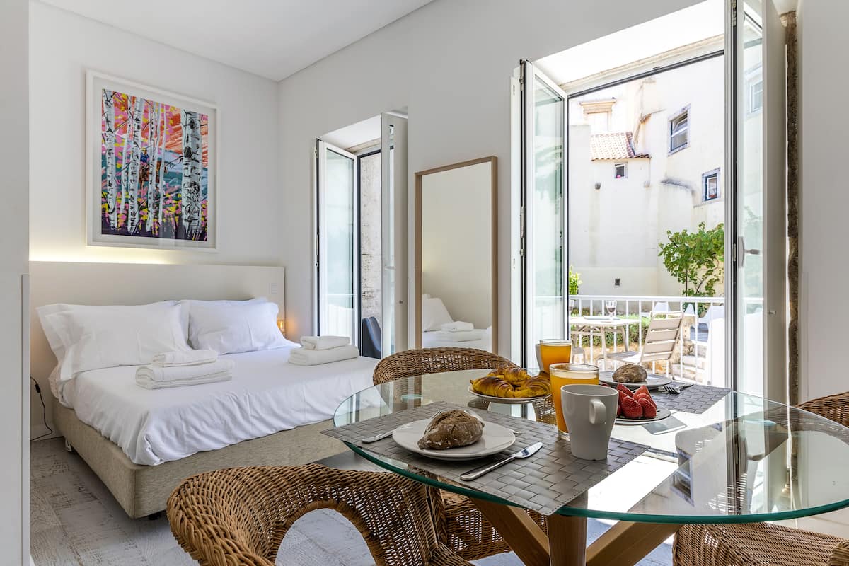 A hotel room with a bed, a glass table with breakfast, a wicker chair, and a balcony with a view.