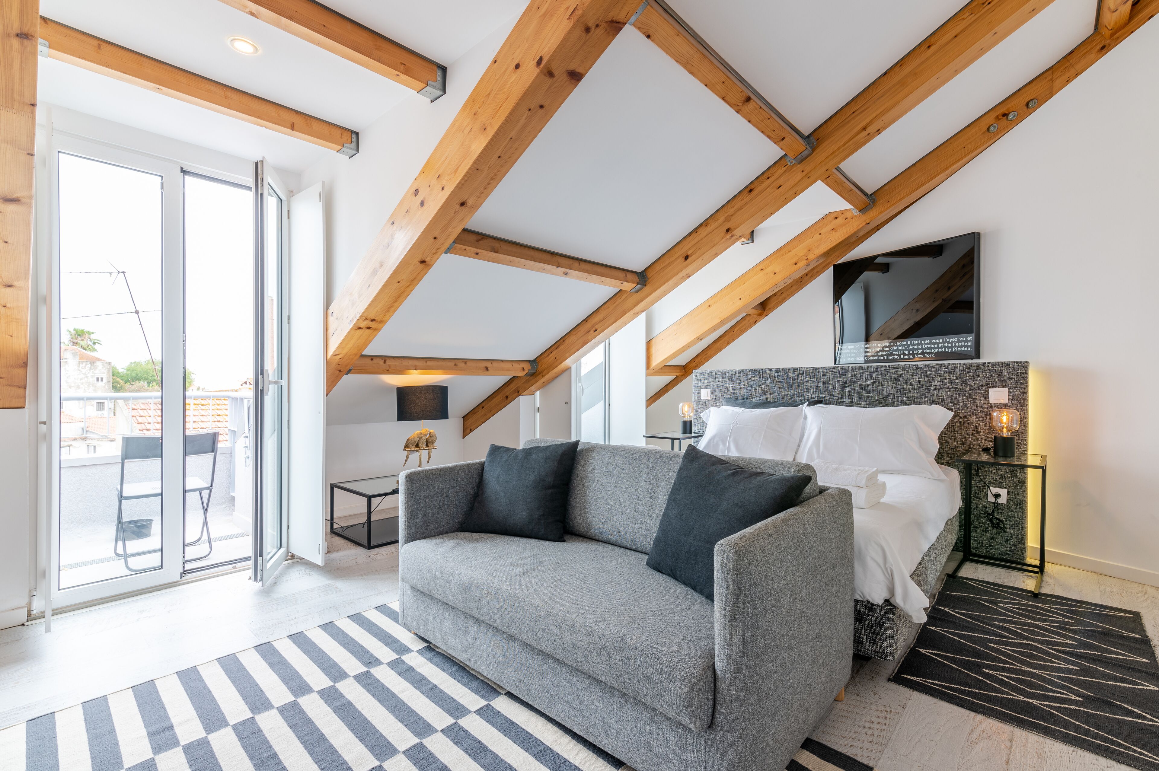 Un dormitorio moderno con techo inclinado, un sofá gris, una cama con ropa de cama blanca y una mesita con una lámpara.