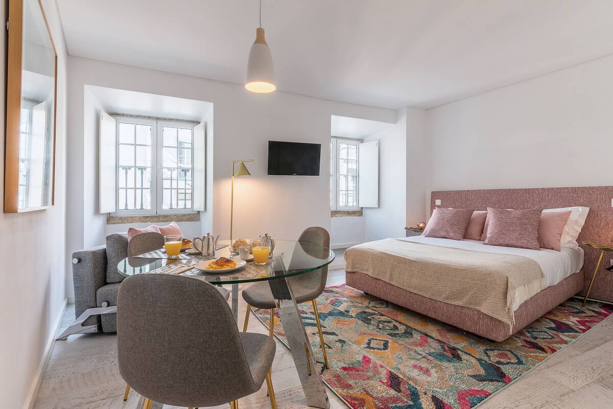 A modern hotel room with a glass dining table, a bed with pink bedding, and a colorful rug.
