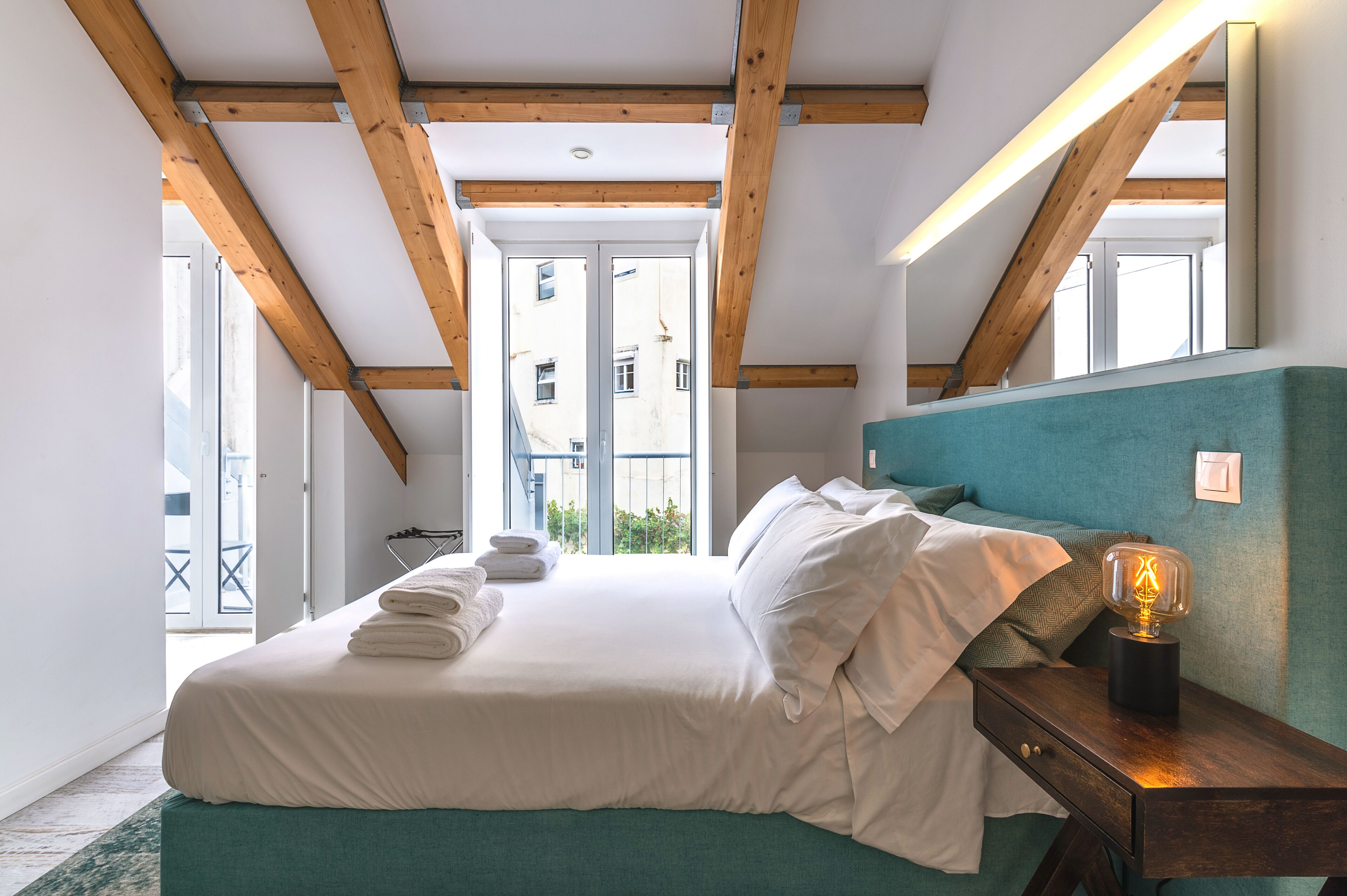 Une chambre avec un grand lit, des poutres en bois, une tête de lit de couleur turquoise et une lampe de chevet.