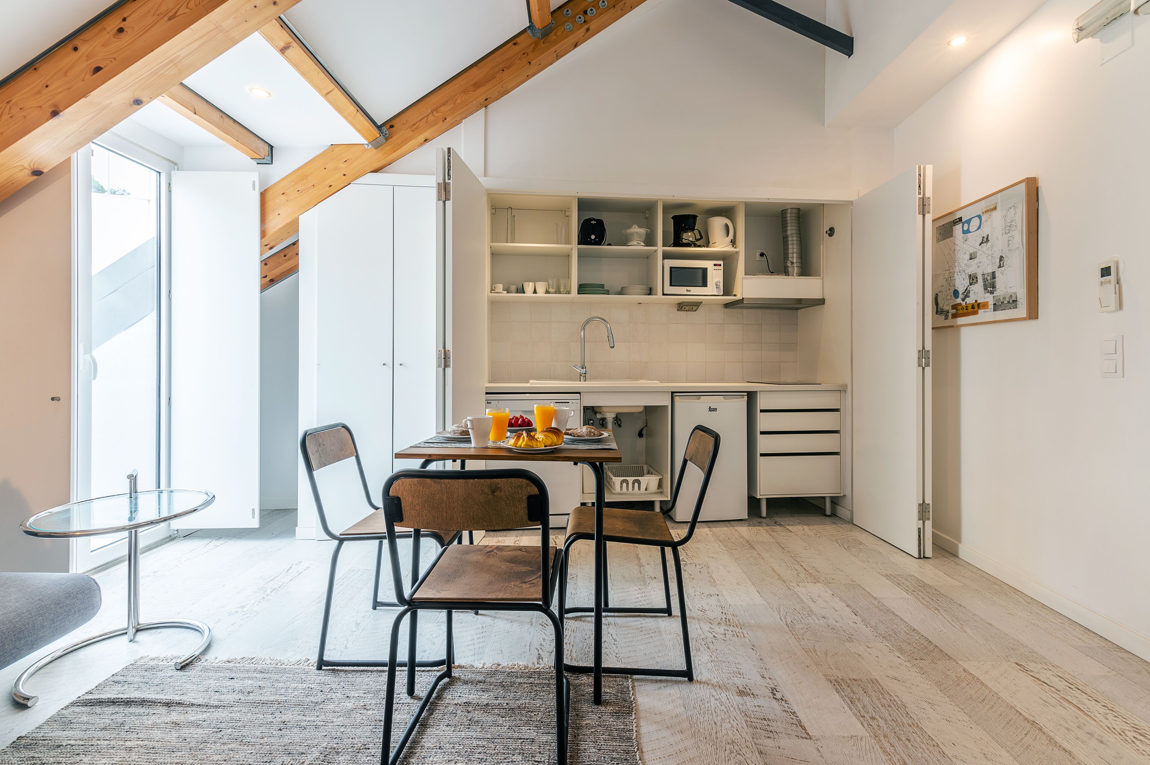 Cocina compacta con armarios blancos, microondas y un lavabo. Hay una mesa de comedor con sillas y una pequeña mesa de vidrio. La habitación tiene vigas de madera en el techo y un ventanal grande.