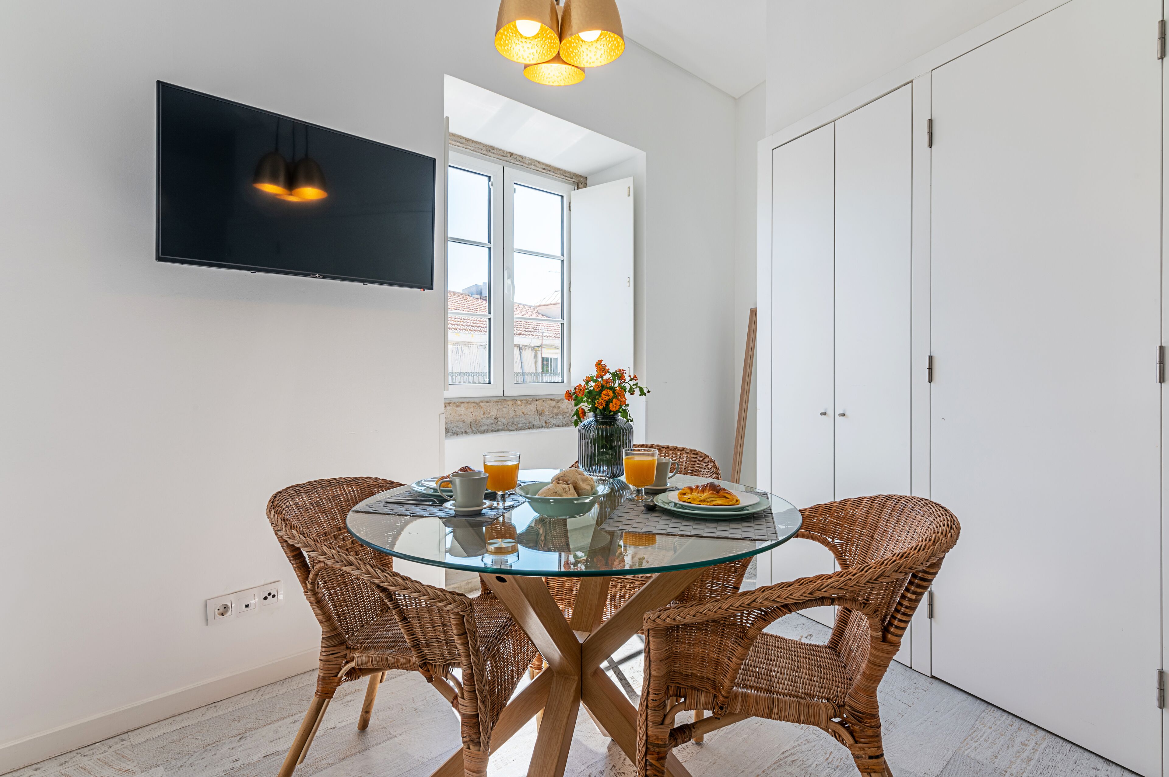 A dining area with a glass table surrounded by wicker chairs, a flat-screen TV mounted on the wall, and a window with a view of buildings.