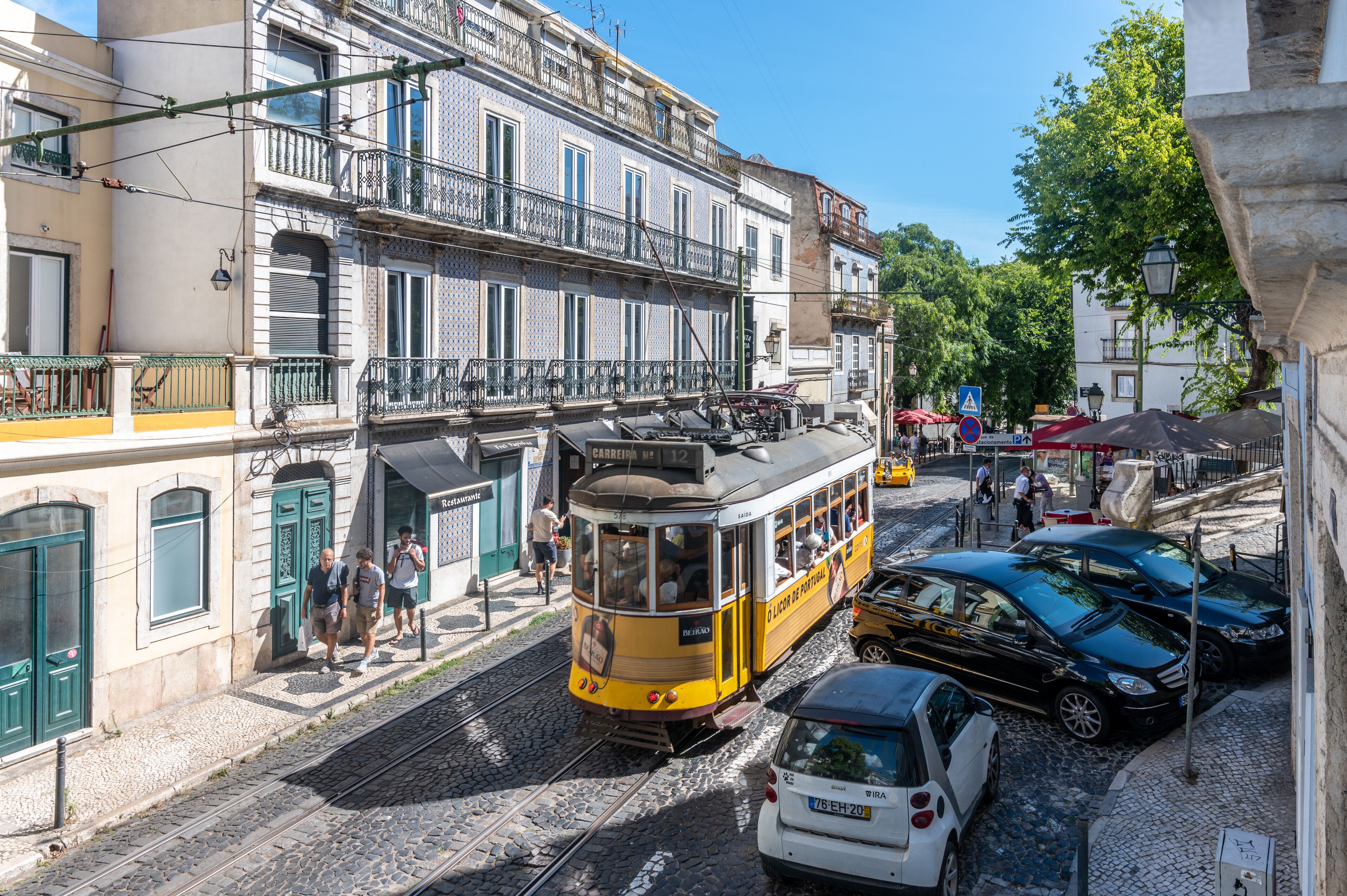 Un tramway sur les rails, des voitures garées et des piétons dans une rue pavée d’une ville historique.