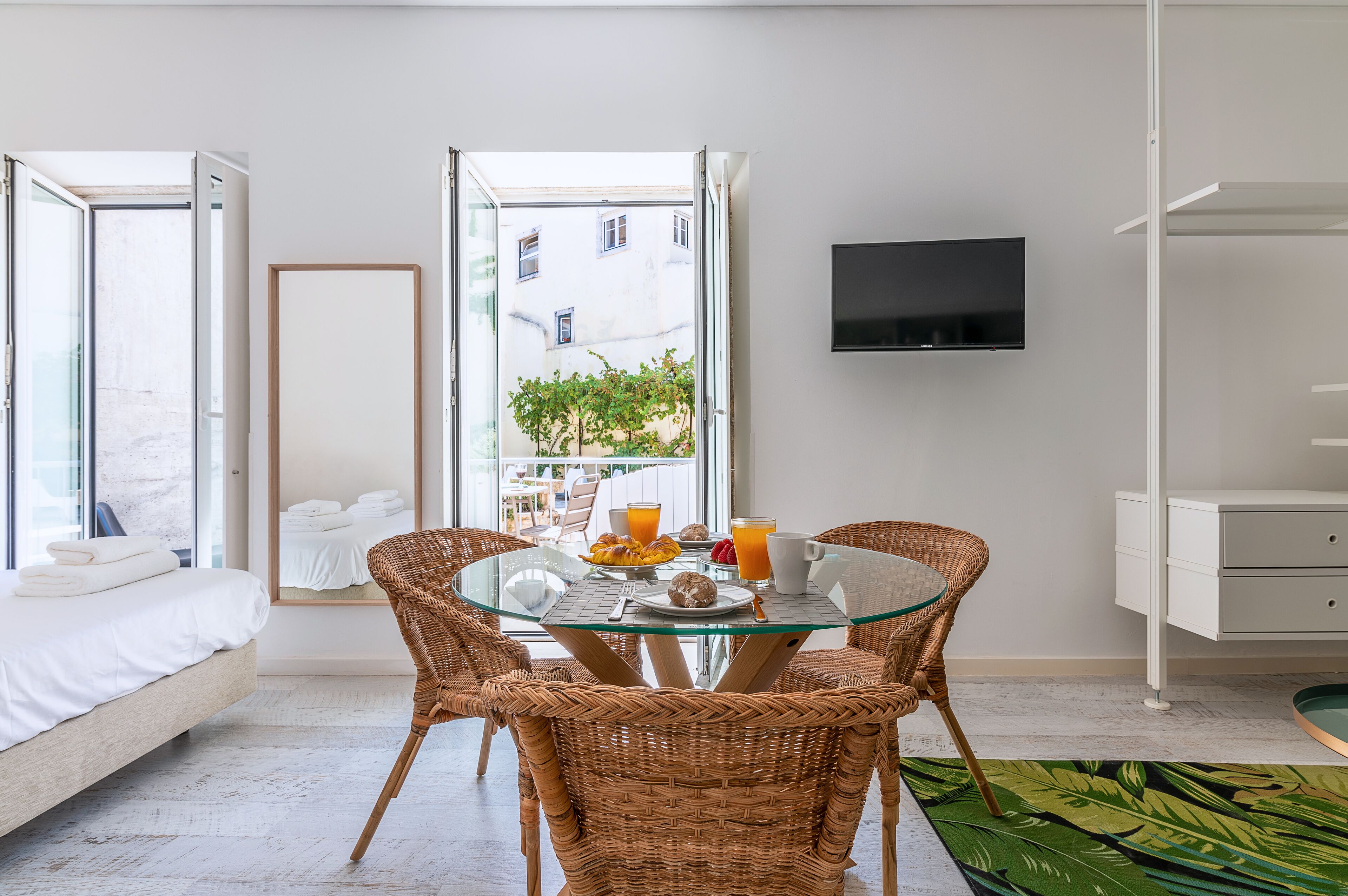 Une chambre d’hôtel moderne avec une table à manger en verre, des chaises en osier, un lit, une télévision et un balcon offrant une vue sur la ville.