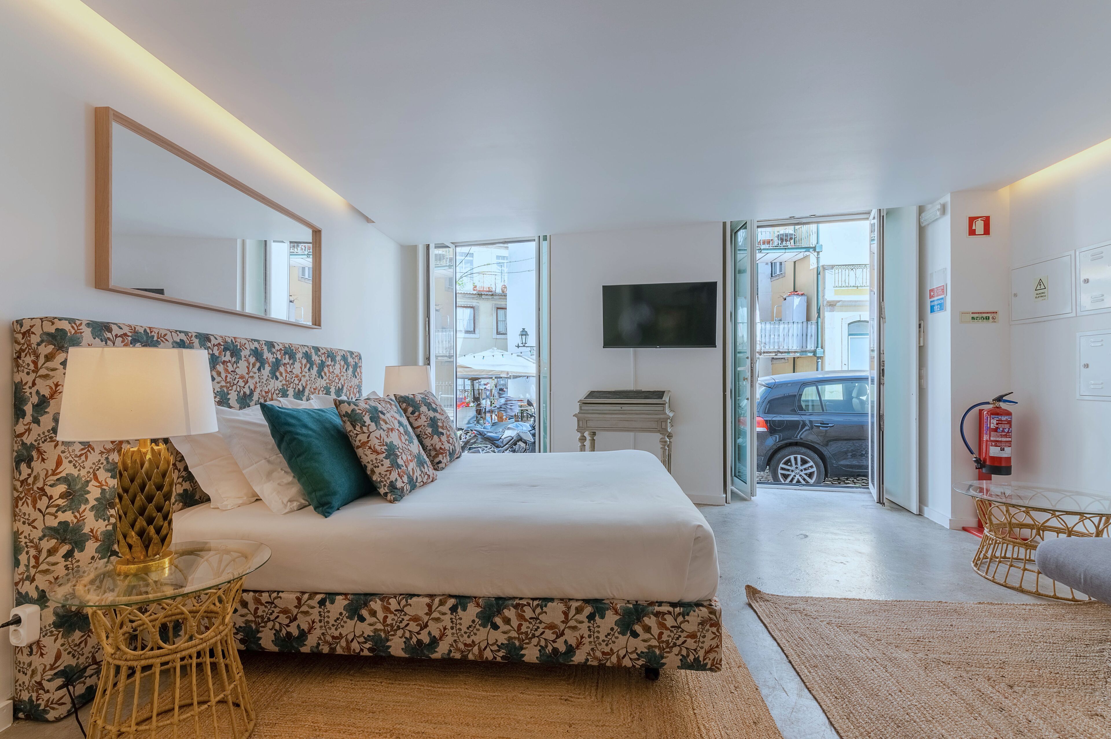 A hotel room with a bed, a TV, a mirror, and a view of the street outside.