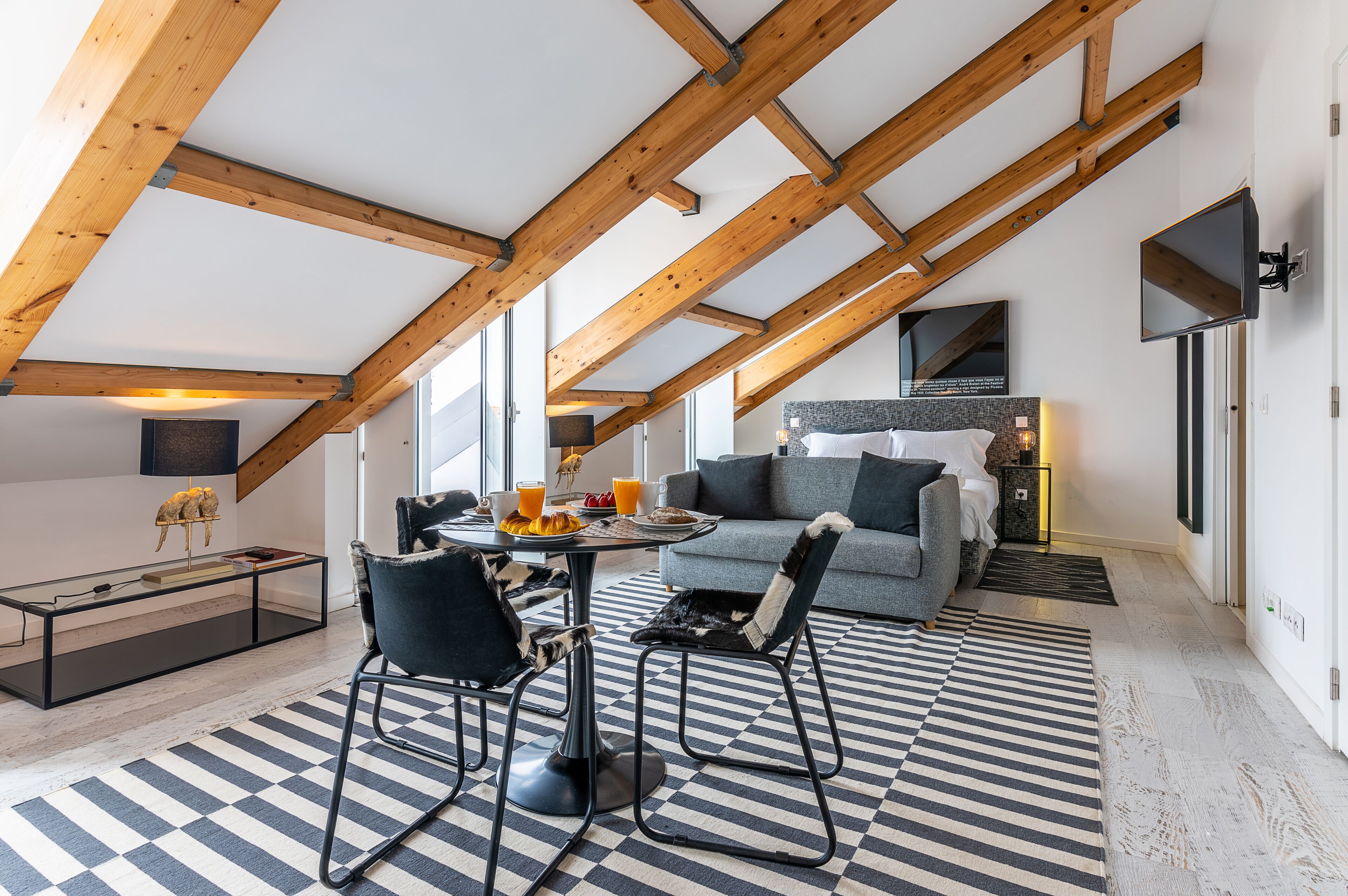 A loft-style bedroom with exposed wooden beams, a checkered floor, a bed with a gray headboard, and a television mounted on the wall.
