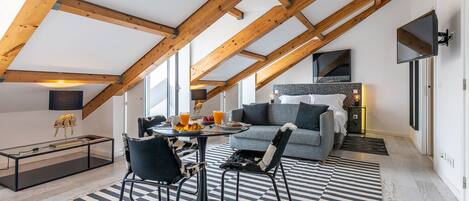 A loft-style bedroom with exposed wooden beams, a checkered floor, a bed with a gray headboard, and a television mounted on the wall.