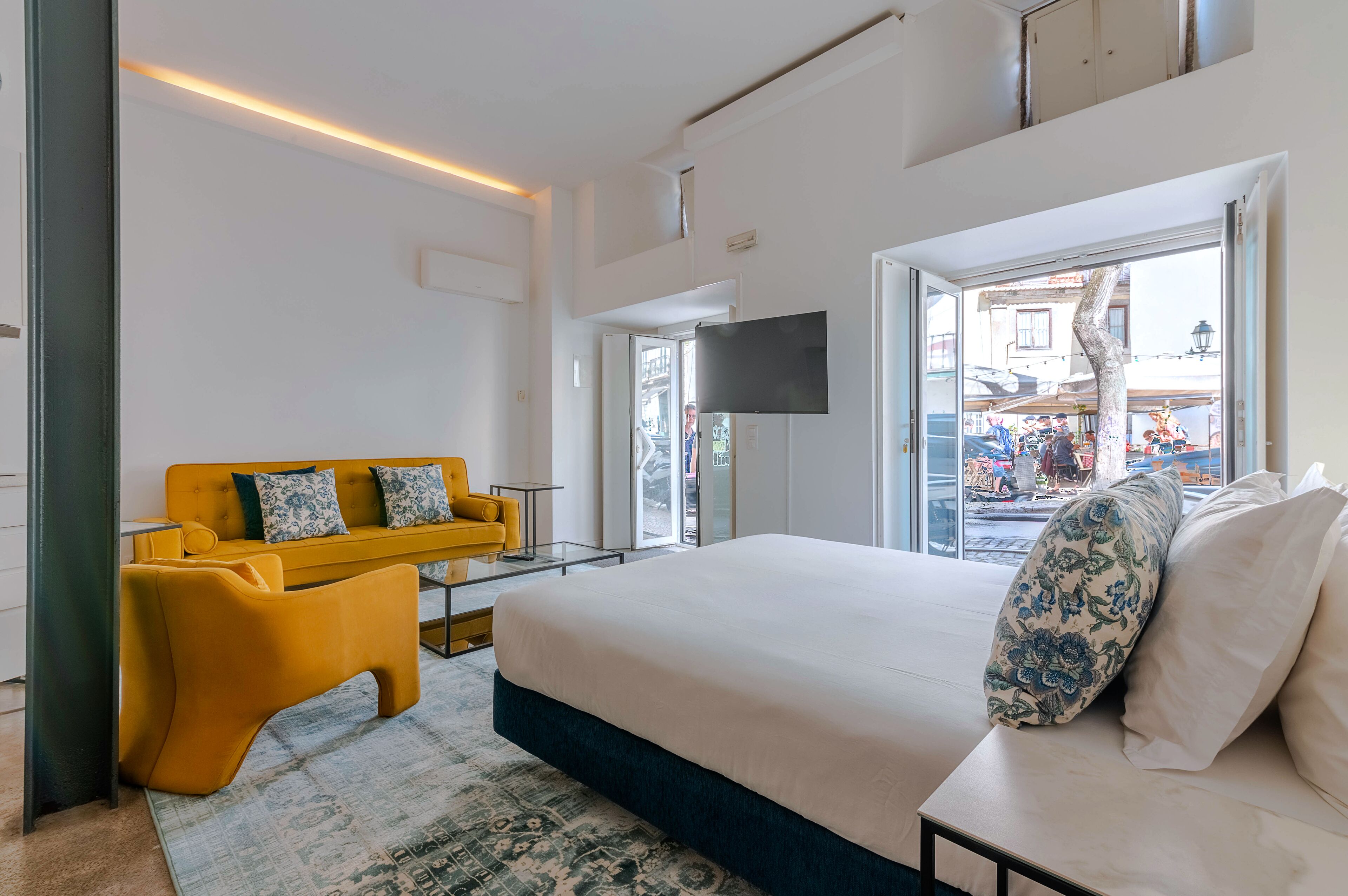 A modern hotel room with a large bed, a yellow sofa, a flat-screen TV, and a view of the street outside.
