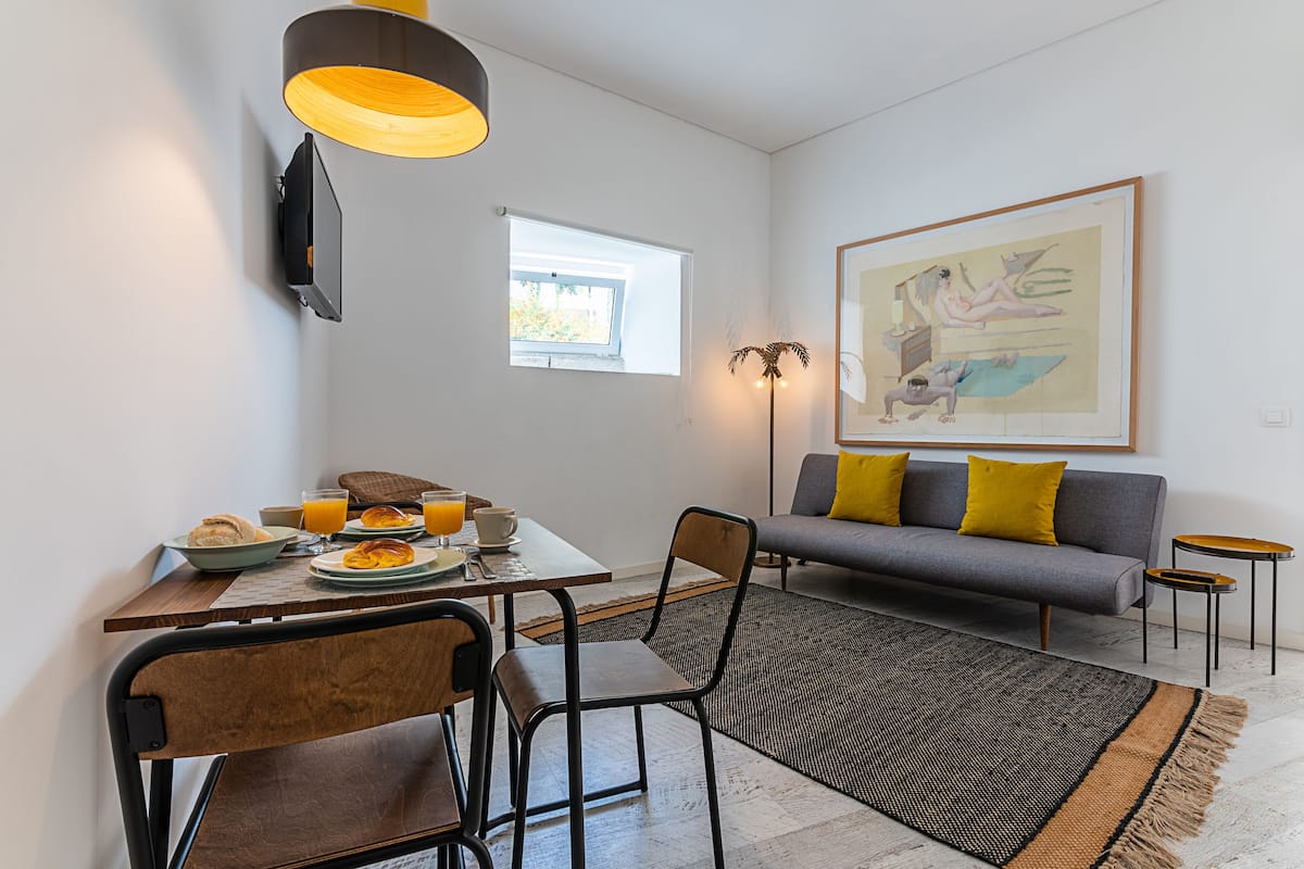 A modern living room with a grey sofa, yellow cushions, a dining table with breakfast, and a large painting on the wall.