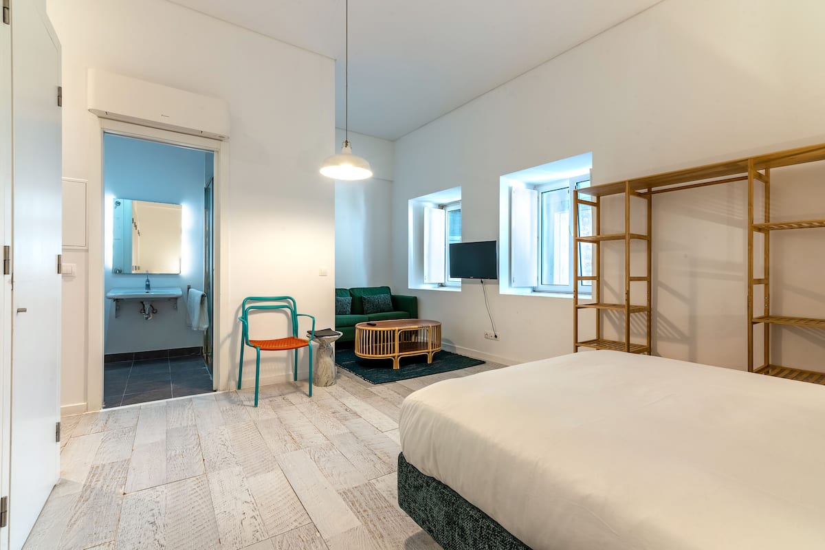 A modern hotel room with a large bed, a green chair, a small table, a TV, and a window with a view.