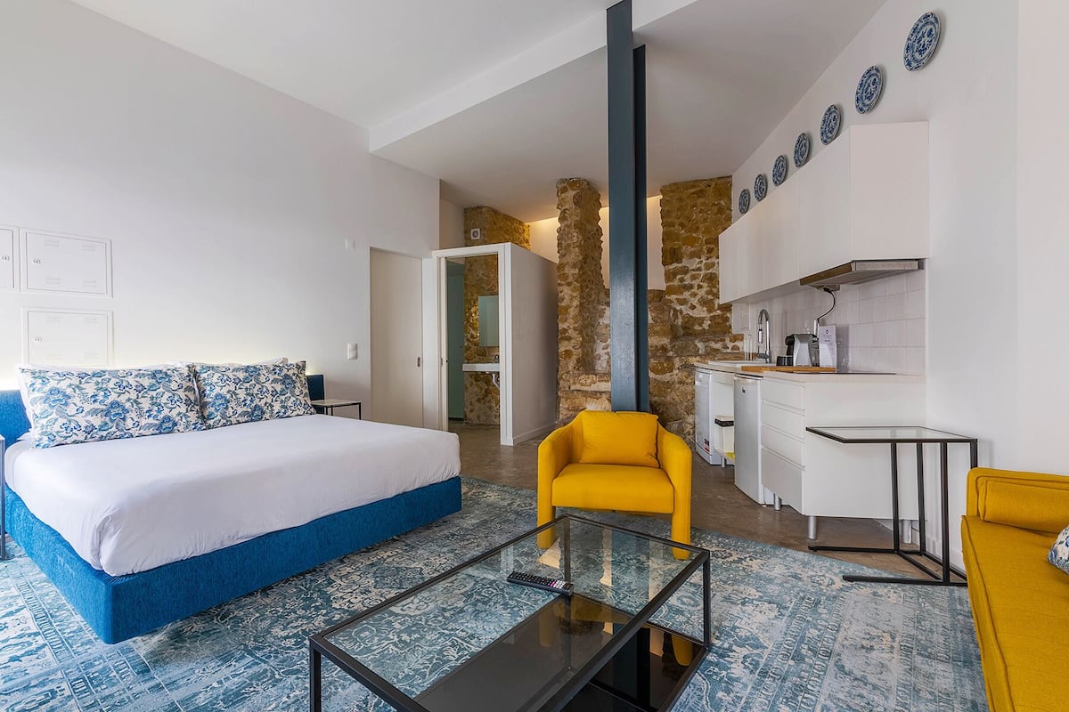 A bedroom with a bed, a yellow armchair, a glass coffee table, and a kitchenette in the background.