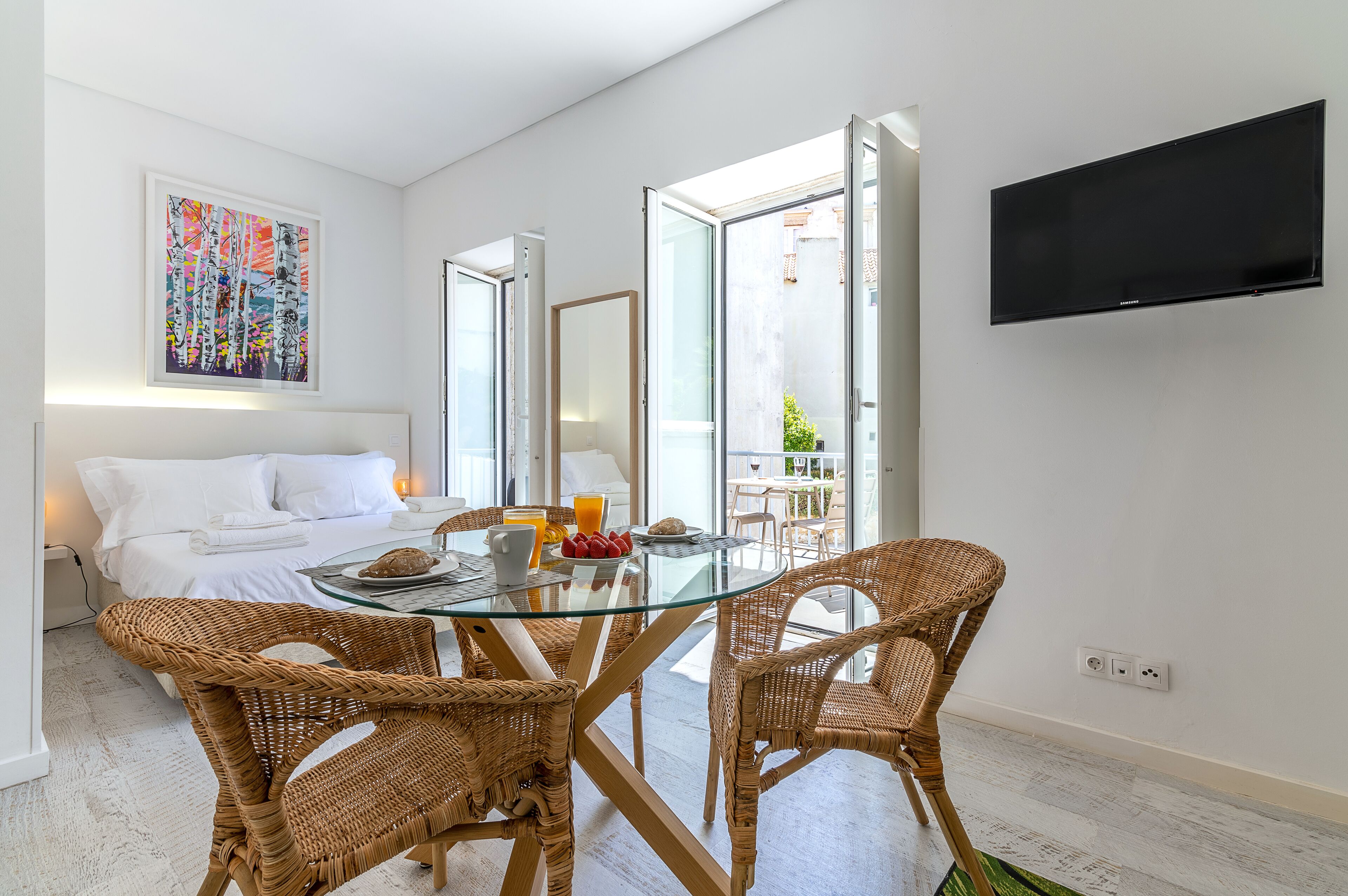 A modern hotel room with a glass dining table, wicker chairs, a bed with white linens, a painting on the wall, and a TV mounted on the wall.