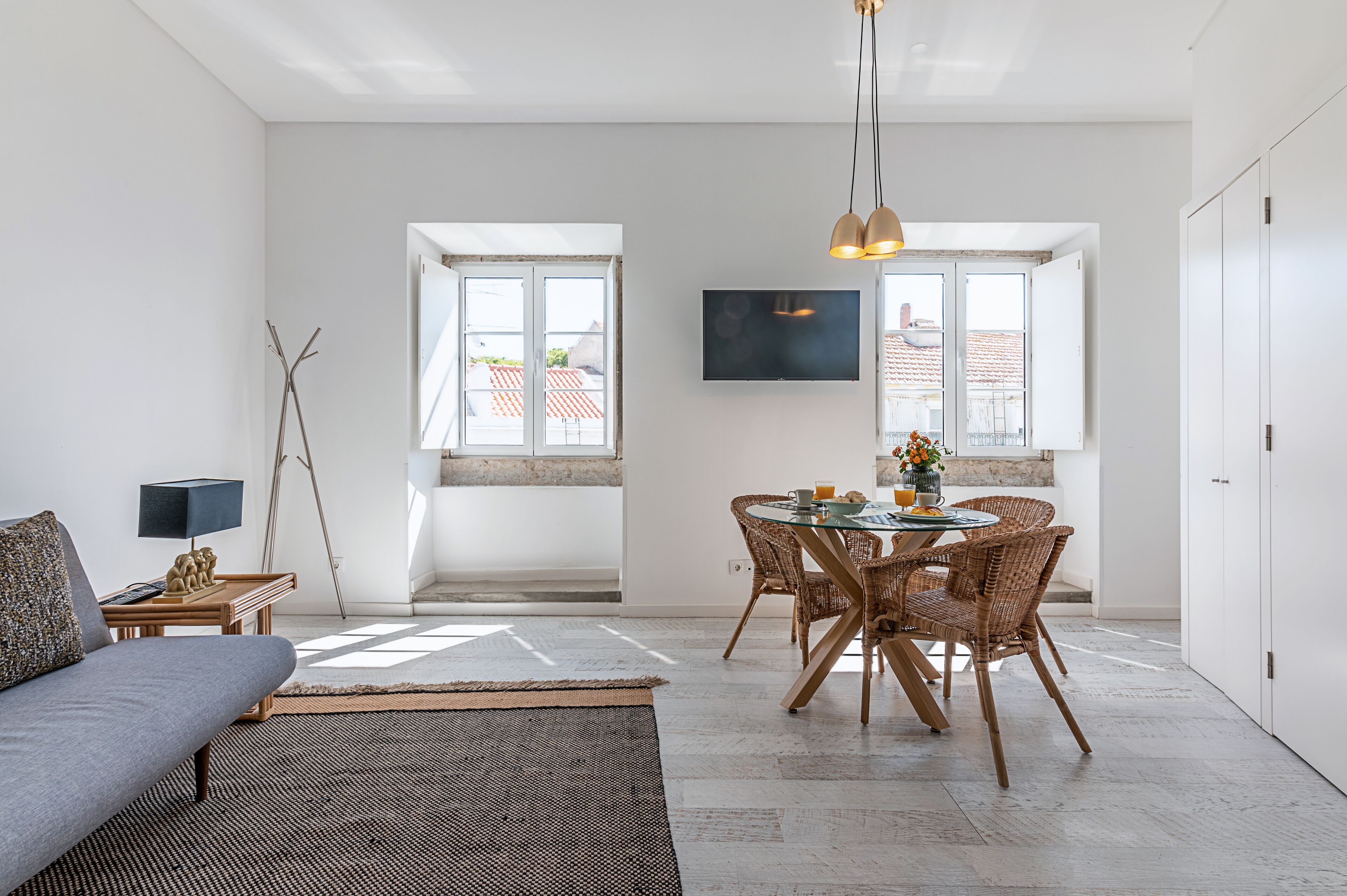A modern living room with a sofa, a dining table with wicker chairs, a TV mounted on the wall, and large windows with a view of buildings outside.
