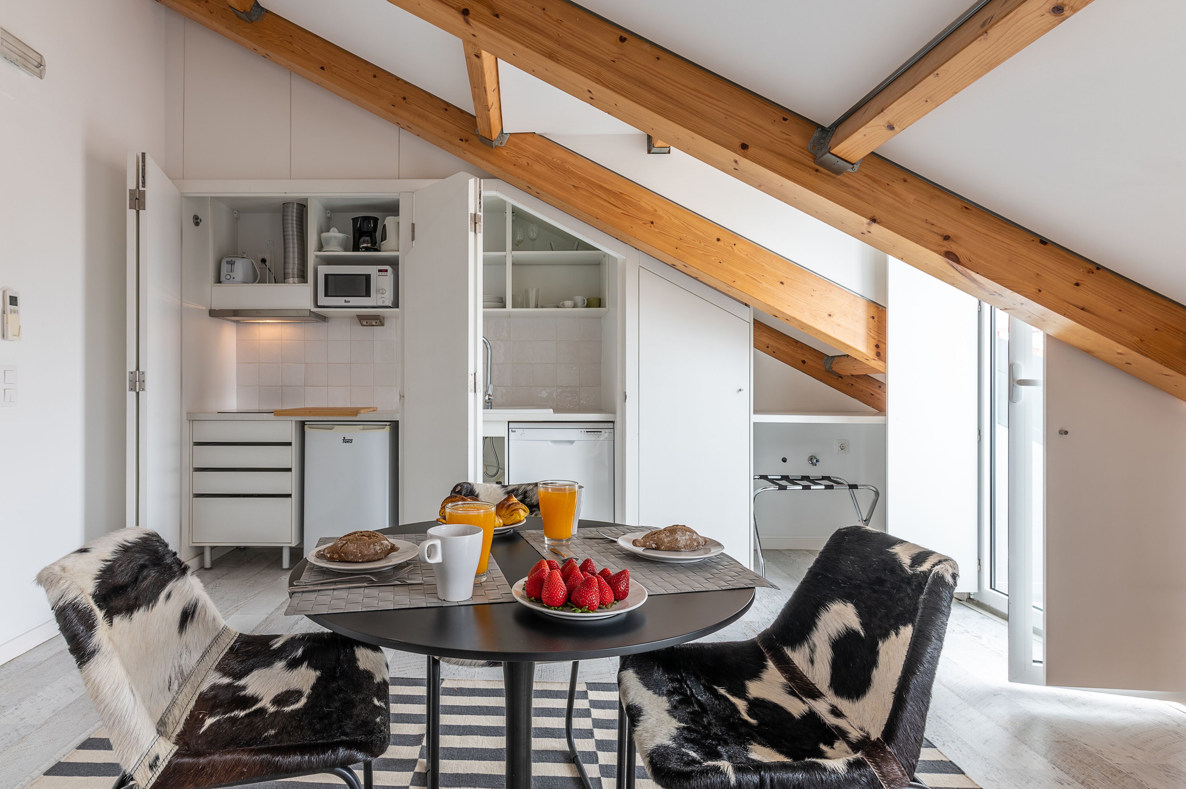 A modern kitchen with white cabinets, a microwave, and a coffee maker. There is a dining table with a black tablecloth, two chairs with black and white fur upholstery, and a plate of strawberries and a croissant.