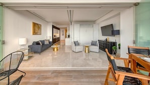 Una sala moderna con un sofá, una mesa de centro y sillas. Hay un televisor montado en la pared y una puerta que da a otra habitación.