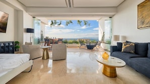 Una sala moderna con cama, sofá, mesa de centro y sillas. Hay un ventanal con vistas al exterior.