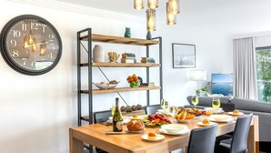 Un coin repas avec une table en bois mise pour le repas, une bibliothèque agrémentée d’objets décoratifs et une horloge murale.