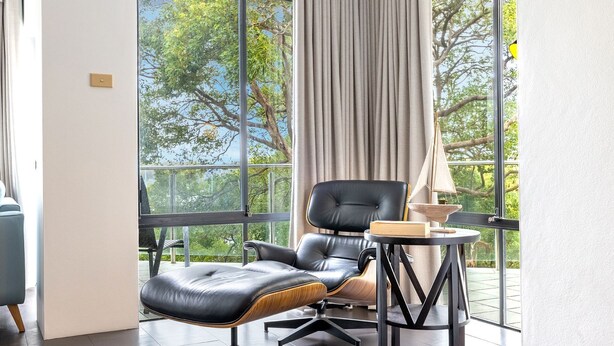 Une chambre moderne avec un fauteuil en cuir, une petite table et une vue sur les arbres à travers de grandes fenêtres.
