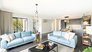 Un salon moderne avec deux canapés bleu clair, une table basse sur laquelle sont disposées des pâtisseries, et un coin cuisine équipé d’un four à micro-ondes et d’un panier de fruits.