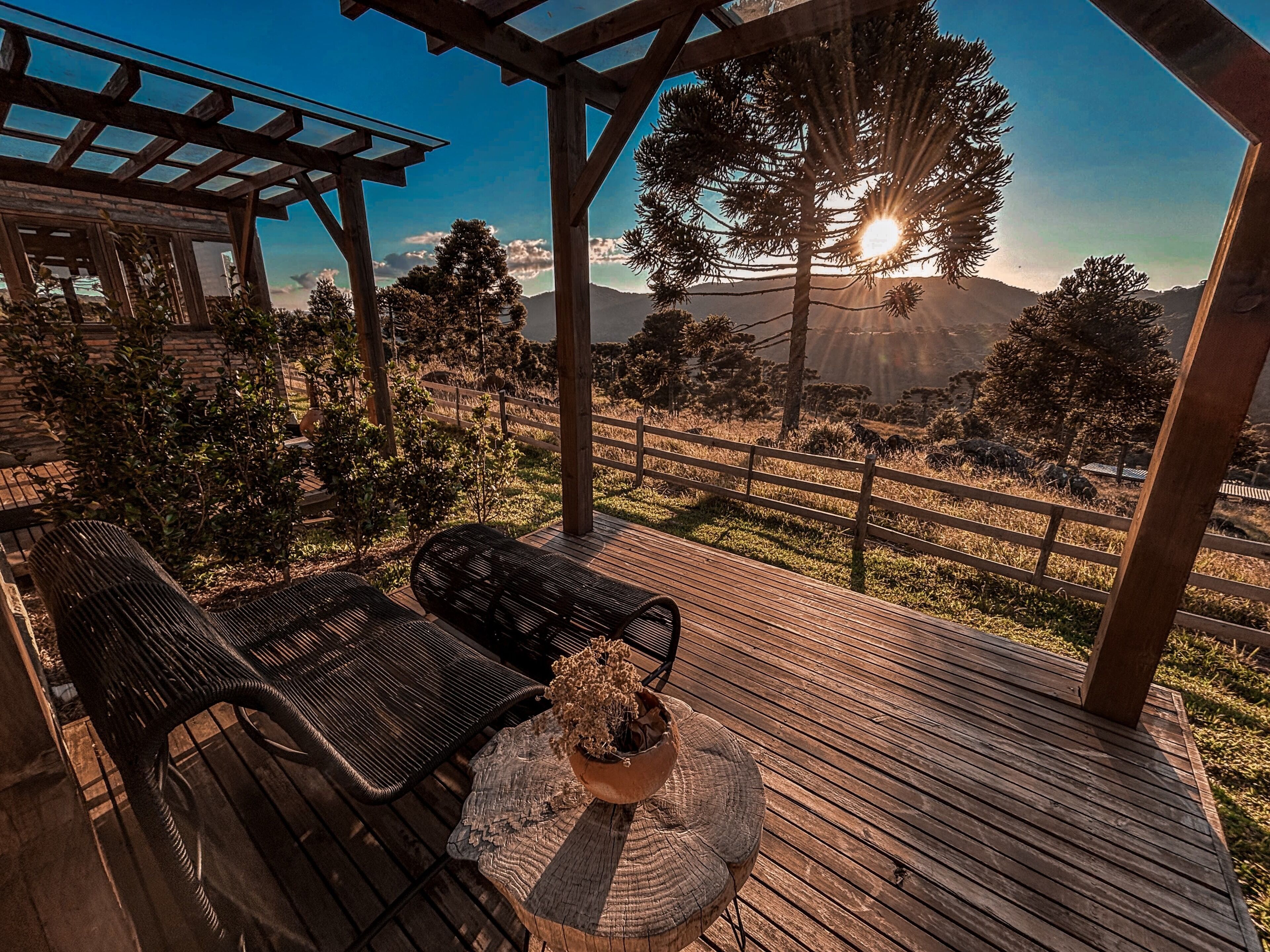 A wooden deck with outdoor furniture, a pergola, and a view of a mountainous landscape during sunset.