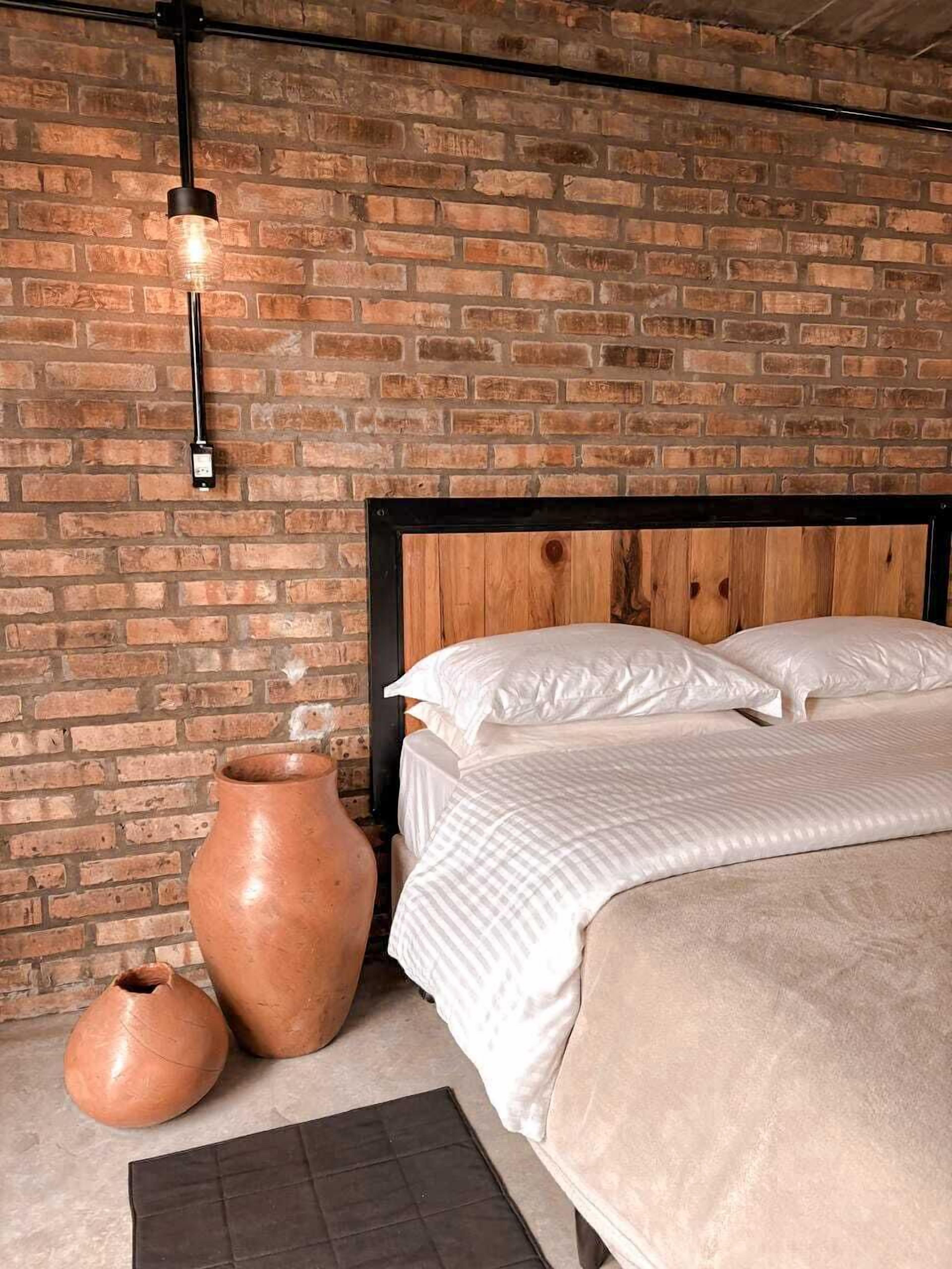 Ein Schlafzimmer mit Backsteinwand, Holzkopfstütze, einem Bett mit weißer Bettwäsche und zwei Terrakotta-Töpfen.