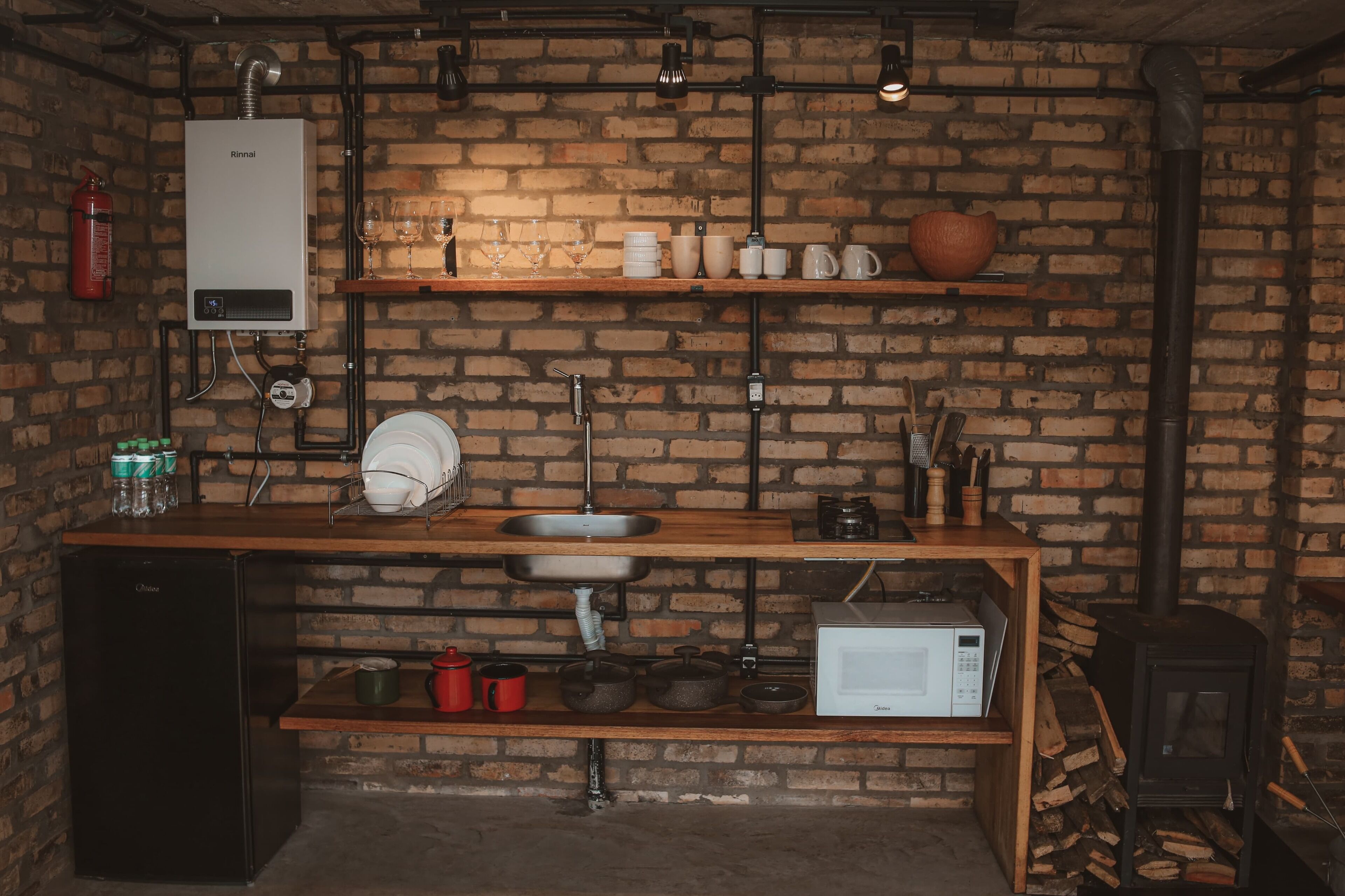 Una cocina con pared de ladrillo, caldera Rinnai, lavamanos, microondas y estufa a leña.