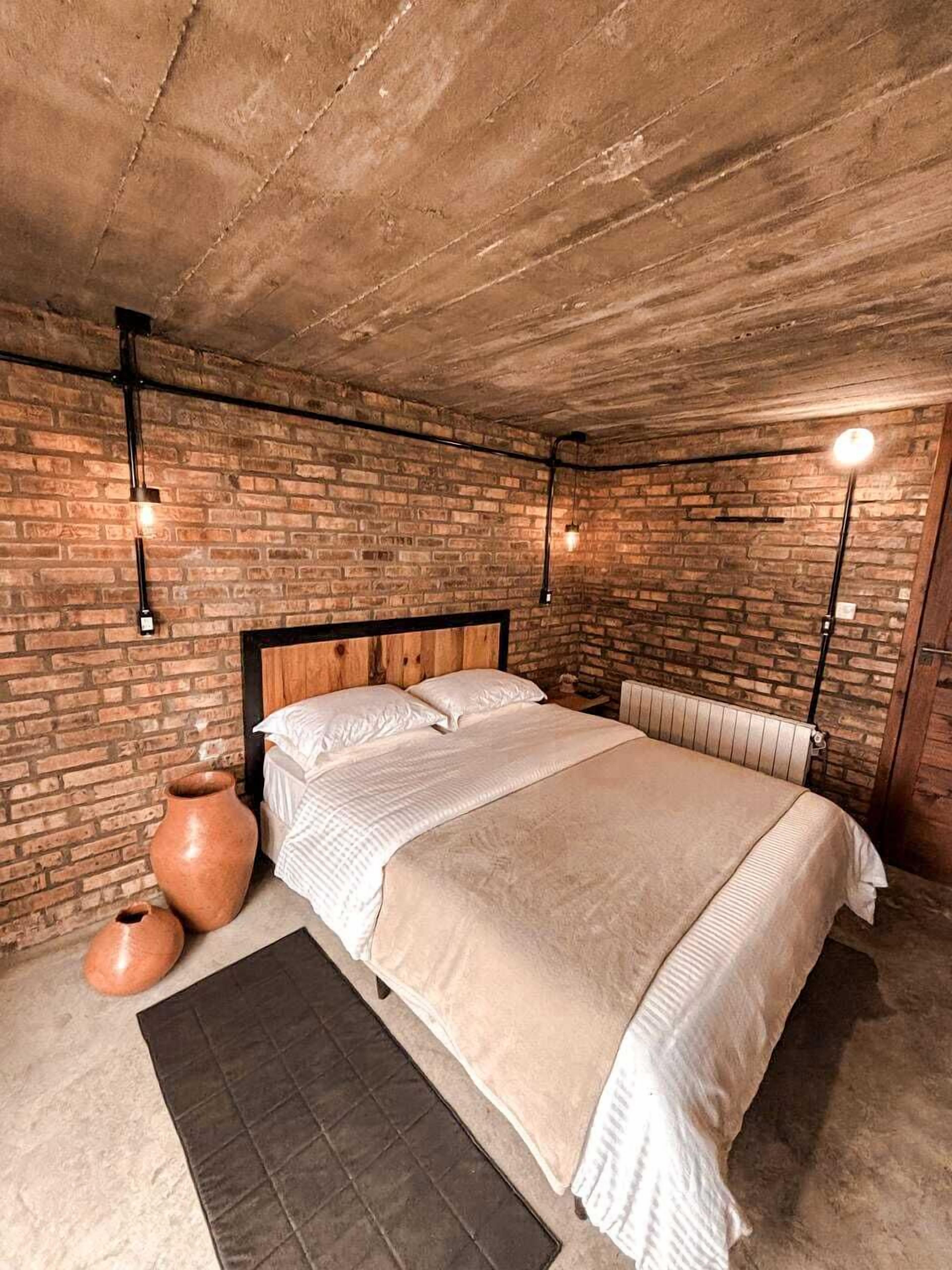 Une chambre avec un mur en briques, une tête de lit en bois, un lit avec une literie blanche, deux lampes fixées au mur et un radiateur.