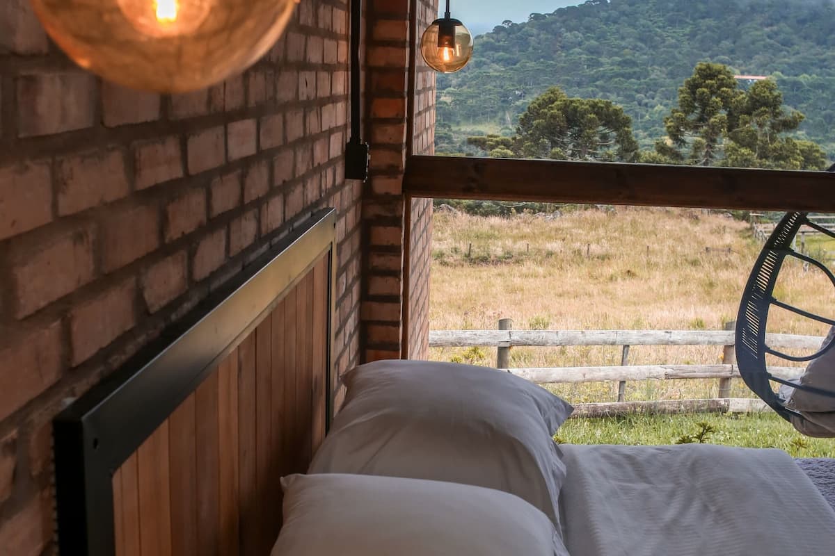 Ein Bett mit weißer Bettwäsche in einem Zimmer mit freiliegenden Backsteinwänden und Blick auf ein Wiesenfeld und Berge.