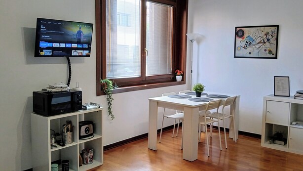 A room with a TV on the wall, a dining table with chairs, a white cabinet with shelves, a window with blinds, and a painting on the wall. - Mi casa es tu casa PN.Free Parking. Great location. (Pordenone)