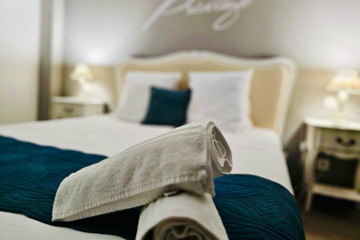 Een net opgemaakt bed met een opgerolde handdoek en een blauwgroene deken, een bedlamp en een muur met de tekst 'Pavilion' in sierletters.