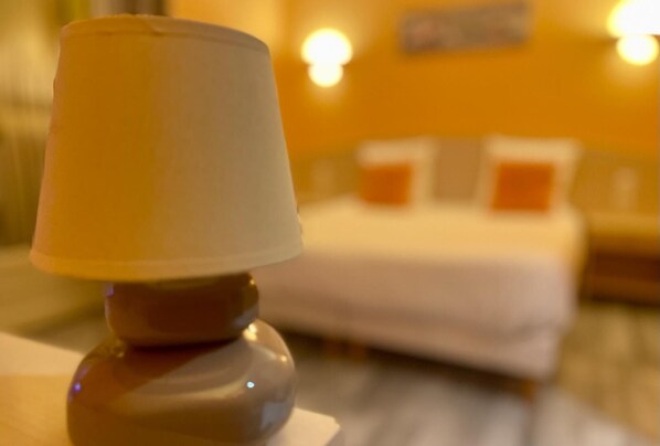 Un gros plan d’une lampe posée sur une table, avec un arrière-plan flou représentant un lit et des appliques murales.