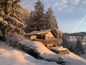 Eine Chalet mit einem hölzernen Balkon, umgeben von schneebedeckten Bäumen und einer Steinmauer.