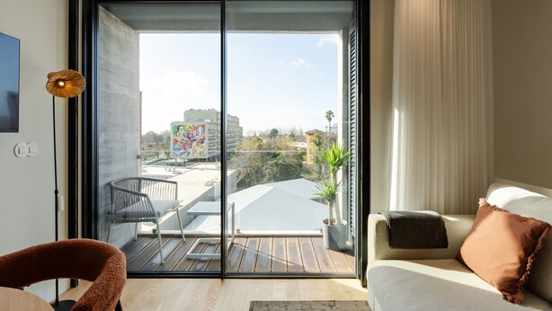 A modern living room with a sliding glass door leading to a balcony, a sofa with cushions, a chair, and a potted plant. - Feel Porto Corporate Housing Boavista (Porto)