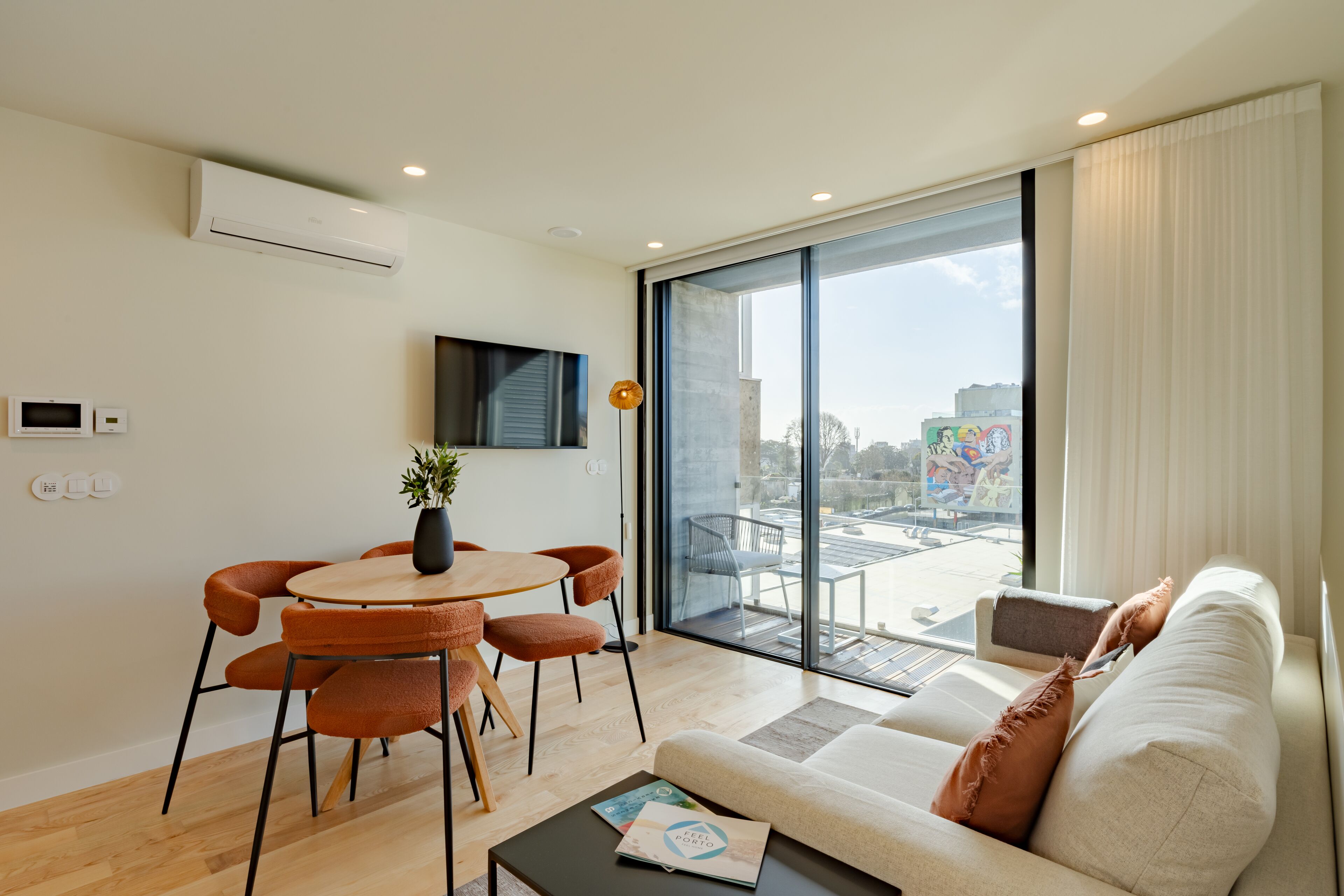 A modern living room with a sofa, a dining table with chairs, a TV mounted on the wall, and a large window offering a city view.