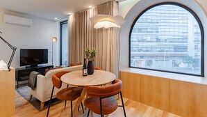 A modern living room with a round wooden table, orange chairs, a TV, and a large window with a view of a building. - Feel Porto Corporate Housing Boavista (Porto)