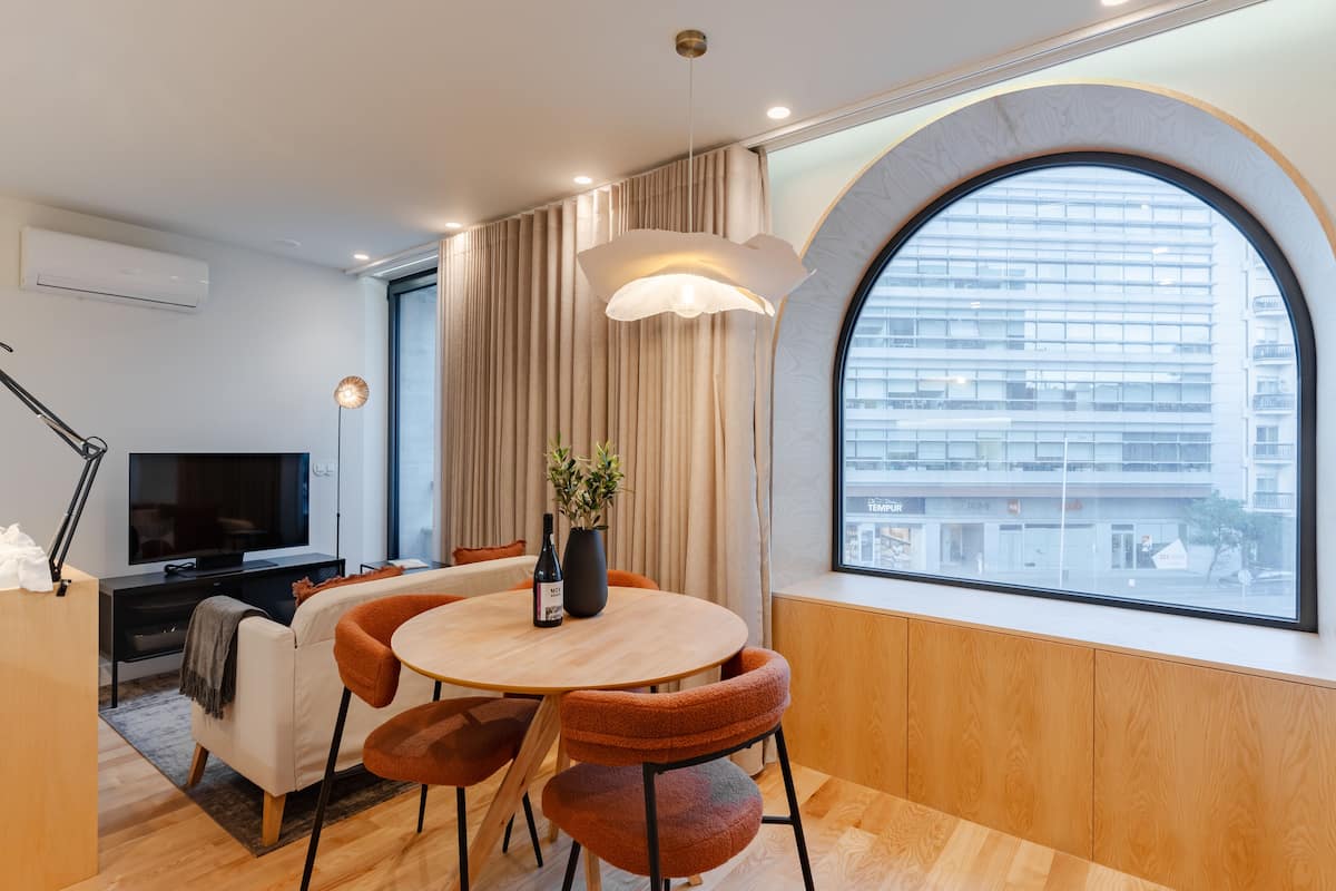 Una sala de estar moderna con una mesa redonda de madera, sillas naranjas, un televisor y un ventanal con vistas a un edificio.