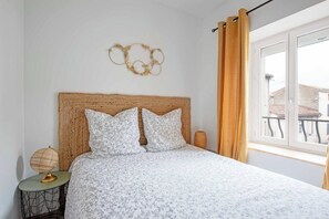 Et soveværelse med en vævet sengegavl, blomstret sengetøj, et natbord med lampe og et vindue med gardiner.