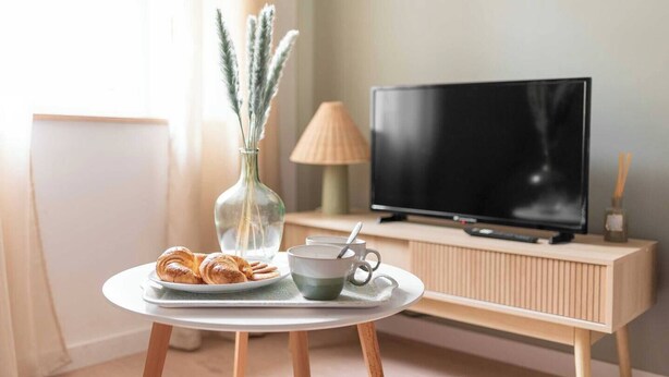 A cozy living room with a round table, a plate of croissants, a cup, a vase with dried flowers, a lamp, and a TV on a wooden stand.