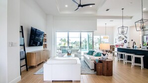 A spacious living area with a large window, a ceiling fan, and a modern kitchen with white cabinetry and bar stools. - Luxury Waterfront 4BR Home w/ Pool & Hot Tub (Cape Coral)