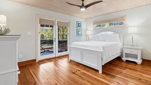 Une chambre avec un ventilateur de plafond en bois, un lit avec une literie blanche, une table de chevet avec une lampe, et une vue sur un espace extérieur, visible à travers une porte vitrée.