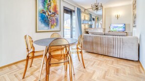 Een moderne woonkamer met een ronde eettafel, transparante stoelen, een bank, een televisie en een groot abstract schilderij.