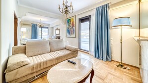 Een woonkamer met een beige bank, een ronde salontafel, een kroonluchter en een groot raam met blauwe gordijnen.